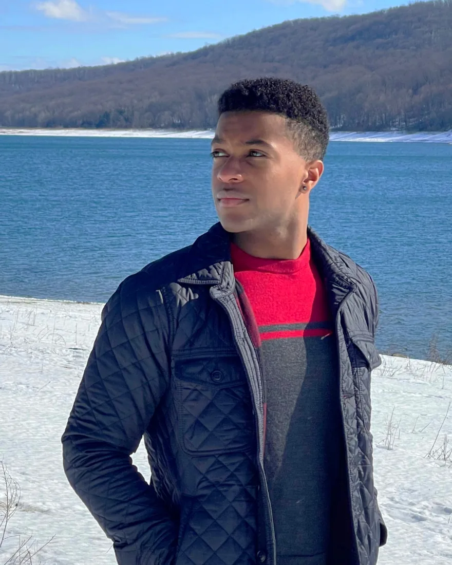 Isaac Mineo standing on a snow-covered bank in a dark quilted jacket, a reservoir and wooded hills behind him.